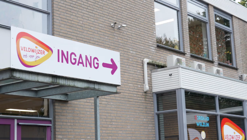 Ingang van het gebouw van De Veldwijzer. Met logo 'Veldwijzer ook voor jou' en logo 'Cordaad welzijn' in beeld.